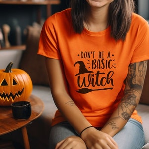 以下が含まれることがあります： オレンジ色のTシャツを着た女性。Tシャツには黒い文字で「Don't be a basic witch」と書かれており、魔女の帽子とほうきのグラフィックが描かれている。彼女はソファに座っており、彼女の前にテーブルがあり、その上にカボチャが置かれている。