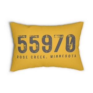 Puede incluir: Cojín rectangular amarillo mostaza con el texto "55970 ROSE CREEK, MINNESOTA" en letras grises desgastadas. Este artículo es un elemento decorativo, ideal para un sofá o una cama.
