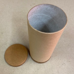 May include: A brown cardboard tube with a lid. The tube is open and the inside is lined with white paper. The lid is off the tube and is sitting on a light brown surface.