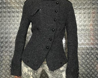 Op de afbeelding: Een donkergrijze wollen jas met een hoge kraag en asymmetrische knoopsluiting. De jas heeft zwarte knopen en decoratieve details op de schouders en mouwen. Het kledingstuk wordt gedragen over een broek met patroon.