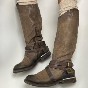 May include: Brown leather knee-high boots with a distressed finish. The boots feature a side zipper, decorative buckles, and straps. The boots have a rugged, worn appearance, suggesting a vintage or pre-loved style.
