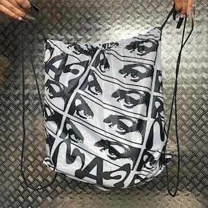 Peut inclure: Sac blanc à cordon de serrage avec un motif d'yeux noirs répétés et le mot "MASK" en grandes lettres noires. Le sac a des cordons noirs.