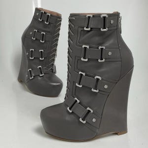 Könnte beinhalten: Graue Keil-Ankle-Boots mit mehreren silbernen Schnallen und Schnürung vorne. Die Stiefel haben eine Plateau-Sohle und einen Reißverschluss hinten. Der Keilabsatz sorgt für einen stilvollen und einzigartigen Look.