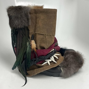 Könnte beinhalten: Ein handgefertigter Stiefel mit einem Obermaterial aus braunem Canvas und einem umgeschlagenen Design. Er verfügt über dunkle Pelzbesätze, Federn und dekorative Elemente, darunter Zähne. Der Stiefel hat eine schwarze Sohle. Das Design deutet auf ein rustikales oder Fantasy-Thema hin.