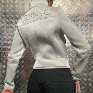 Puede incluir: Una chaqueta corta de color gris claro con cierre de cremallera y un logotipo "Techwear" en relieve en la espalda. La chaqueta tiene una superficie ligeramente texturizada y un ajuste delgado.