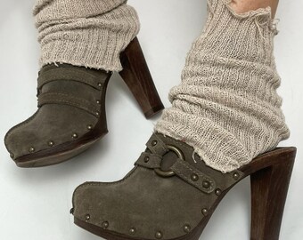 May include: Olive green suede clogs with wooden heels and studded details. The clogs have a buckle closure and are paired with beige knit leg warmers. The heels are approximately 4 inches tall.