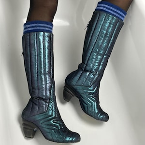 May include: Iridescent teal knee-high boots with a quilted design and a geometric pattern on the toe. The boots have a block heel and a zipper closure. Blue and white striped socks peek out from the top of the boots.