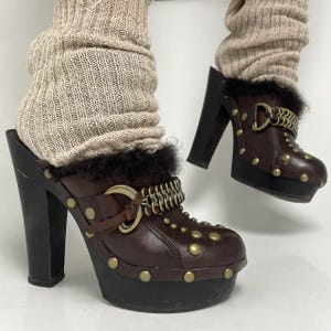 Puede incluir: Zuecos de plataforma de cuero marrón con suela y tacón negros. Los zuecos presentan tachuelas doradas, un detalle de cadena y un ribete de piel. Los tacones miden aproximadamente 10 cm de alto.