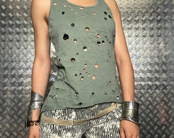 May include: A distressed olive green tank top with numerous circular cutouts. The top is paired with patterned pants and silver bracelets. The overall style suggests a grunge or alternative fashion aesthetic.