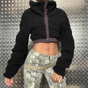 Peut inclure: Veste courte noire avec un col montant et des manches longues. La veste présente un tissu texturé et une bordure violette contrastante le long de la fermeture à boutons. Les manches sont froncées.