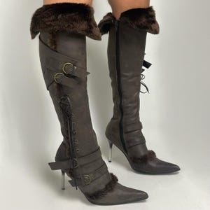 Puede incluir: Botas marrones hasta la rodilla con punta, tacones altos y ribete de piel. Las botas tienen hebillas, correas y una cremallera lateral. Los tacones son de color plateado.
