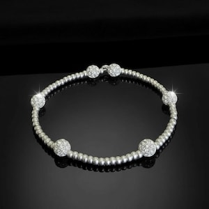 Puede incluir: Pulsera plateada con pequeñas cuentas y seis grandes bolas cubiertas de cristales brillantes. La pulsera se muestra sobre un fondo negro. Una joya delicada. Circunferencia de aproximadamente 18 cm.