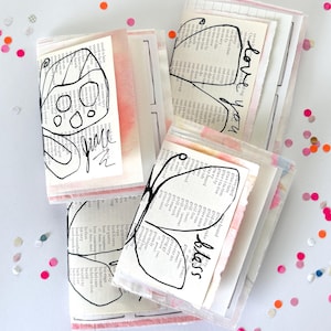 May include: Three small handmade booklets featuring pages from a dictionary with black ink line drawings of butterflies on the covers. The covers include handwritten text that reads "grace," "love you," and "bliss" with soft pink watercolor accents.