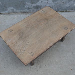 May include: A low rectangular wooden table with a natural light brown finish and visible wood grain. The tabletop features four distinct rectangular joinery details at each corner, connecting to short, sturdy legs beneath the surface.