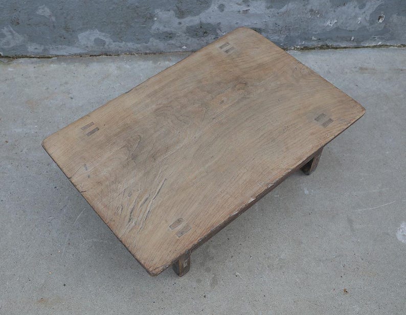 May include: A low rectangular wooden table with a natural light brown finish and visible wood grain. The tabletop features four distinct rectangular joinery details at each corner, connecting to short, sturdy legs beneath the surface.