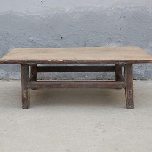 May include: A low rectangular wooden coffee table with a weathered brown finish. The table features a flat, smooth top surface supported by four sturdy legs connected by horizontal wooden stretchers, showing visible signs of age and wear.