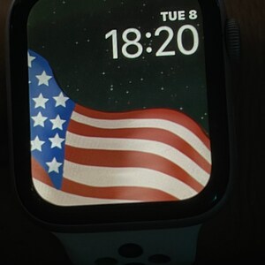 May include: A white Apple Watch with a black face displaying the time 18:20 and the date TUE 8. The watch face has a starry background with a waving American flag in the foreground.