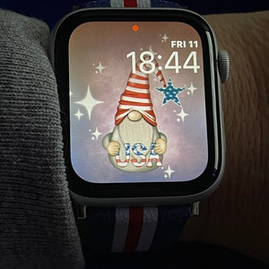 May include: A white Apple Watch with a blue, white, and red striped band. The watch face displays a digital clock with the time 18:44 and the date FRI 11. The background of the watch face is a light pink with a cartoon gnome wearing a red, white, and blue hat and holding a sign that says "USA".