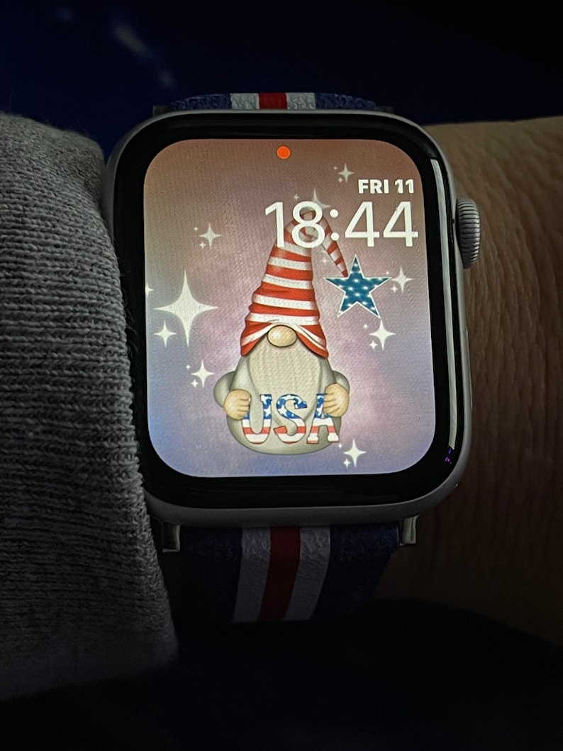 May include: A white Apple Watch with a blue, white, and red striped band. The watch face displays a digital clock with the time 18:44 and the date FRI 11. The background of the watch face is a light pink with a cartoon gnome wearing a red, white, and blue hat and holding a sign that says "USA".