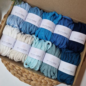 Può includere: Una scatola piena di filato iCord in varie tonalità di blu e crema. Ogni gomitolo è avvolto con un'etichetta che recita "MY LITTLE MIMI". Il filato è ordinatamente disposto in una scatola di cartone, suggerendo un kit di forniture artigianali.