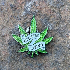 May include: A green glitter marijuana leaf pin with a white banner that reads "Assisted Living".