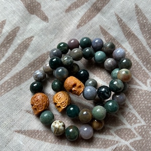 Puede incluir: Dos pulseras hechas de piedras verdes, marrones y grises con tres cuentas de madera talladas en forma de calavera.