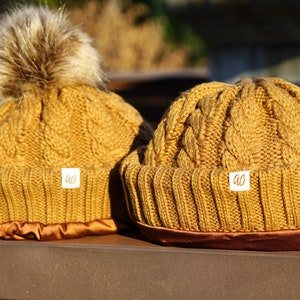 Puede incluir: Dos gorros de punto marrón con un patrón de punto de cable. Un gorro tiene un pompón de piel sintética marrón en la parte superior. Ambos gorros tienen un ribete de cinta de satén marrón y una etiqueta blanca con las letras "AO" en ellos.