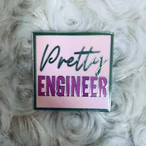 Peut inclure: Une épingle en émail rose et verte avec le texte "Pretty Engineer" en lettres blanches et violettes.