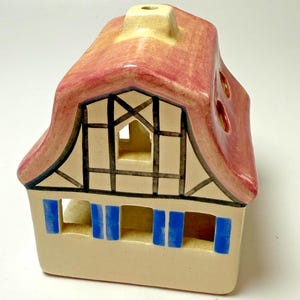 May include: A ceramic house-shaped candle holder with a red roof and beige walls. The house features blue shutters, black and white trim, and a chimney. The roof has three round openings. The candle holder is designed to hold a tea light.