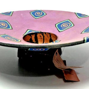 May include: A ceramic serving dish with a pink base and a colorful geometric pattern. The dish has a central hole and a matching base.
