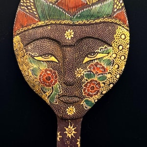 May include: A hand mirror with a dark brown wooden frame shaped like a stylized face. The surface features intricate painted details in yellow, green, and red, including floral patterns on the cheeks, sun motifs on the handle, and a crown design.