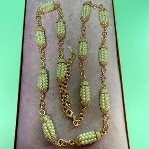 Op de afbeelding: Een goudkleurige ketting met imitatieparels en decoratieve gouden accenten. De ketting wordt gepresenteerd in een roze fluwelen doos met een gouden rand. De ketting heeft cilindrische parelclusters die zijn verbonden door gouden schakels.