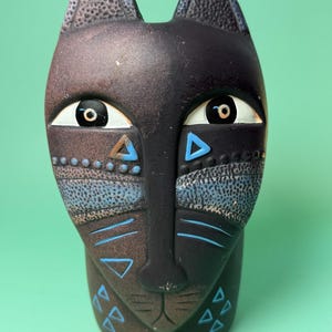 Può includere: Statuetta decorativa a forma di gatto con un design a volto diviso. Un lato è marrone, l'altro nero. Il gatto ha grandi occhi, forme geometriche blu e orecchie strutturate. La statuetta è su uno sfondo verde.
