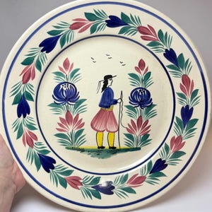 May include: A round ceramic plate with a cream-colored base featuring a hand-painted design of a person in a blue top, pink skirt, and yellow leggings holding a staff. The center is flanked by large blue and pink flowers, with a floral border.