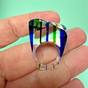 May include: A clear, chunky ring with a rectangular top. The top features vertical stripes of blue, green, and white. The ring is held in a hand against a light green background. The ring's band has a clear, transparent appearance.