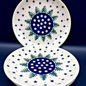 May include: Two round ceramic plates featuring a central dark blue circle decorated with white dots. Surrounding the center are green pine tree motifs and scattered small dark blue stars on a cream-colored background, creating a festive pattern.