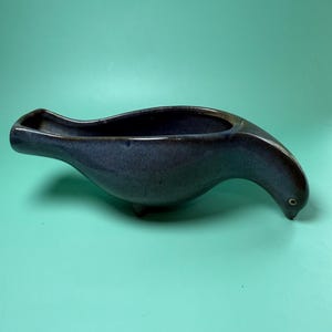 May include: A blue ceramic planter shaped like a seal. The planter has a long, curved body with a small opening at the top. The seal's head is tilted downwards, and it has a small, round eye. The planter is approximately 30 cm long.