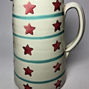 May include: A white ceramic pitcher with a blue and red star pattern. The pitcher has a handle and a wide mouth.