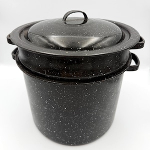 May include: A black enamel stockpot with a white speckled pattern, featuring a matching lid with a central handle and two side handles on the main body. The pot has a cylindrical shape and a smooth, glossy finish throughout its exterior surface.