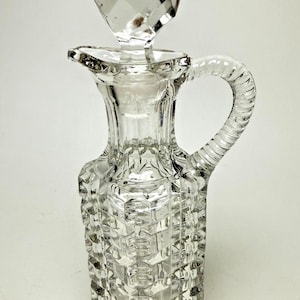 May include: A clear glass cruet with a faceted stopper. The cruet has a square base and a textured, geometric pattern. It features a handle and a spout for pouring. The stopper is diamond-shaped.