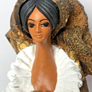 Op de afbeelding: Een keramiek beeldje van een vrouw met zwart haar en een gouden hoofdbedekking. Ze draagt een witte kraag met ruches en heeft rode en gouden accenten op haar borst.