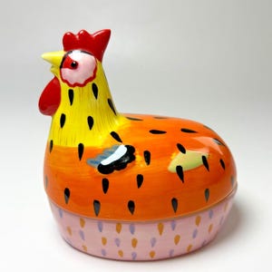May include: A ceramic chicken-shaped container with a bright, hand-painted design. The chicken features a yellow head, orange body, and pink base. Black teardrop shapes are scattered across the body. The comb and wattle are red.
