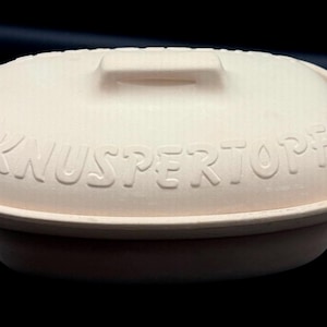 May include: An oval-shaped, light beige ceramic baking dish with a matching lid. The lid features a central handle and the embossed text "KNUSPERTOPF" across the front. The dish has a smooth, matte finish and a simple, functional design.