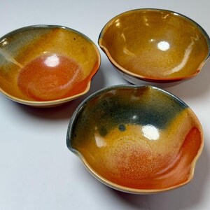 Puede incluir: Tres cuencos de cerámica hechos a mano con una forma orgánica única. Los cuencos presentan una mezcla de colores, incluyendo naranja, amarillo y verde azulado, creando una estética cálida y terrosa. Cada cuenco tiene una pequeña hendidura curva en un lado.
