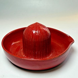 May include: A vibrant red ceramic citrus juicer. The juicer features a central reamer with vertical grooves, surrounded by a bowl with a pouring spout. The glossy finish enhances the rich color of the kitchen tool.