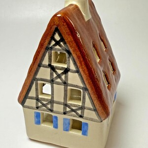 May include: A ceramic house figurine with a brown roof, cream-colored walls, and blue shutters. The roof has cut-out windows. Black lines create a decorative pattern on the walls. A white chimney is on top.