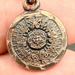 May include: A circular metallic pendant featuring a central relief of a human face surrounded by intricate geometric patterns and symbols. The piece has a dark, oxidized finish with a hammered edge and a small loop at the top for attachment.