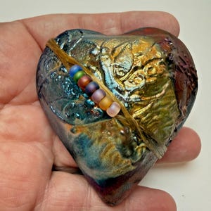 May include: A heart-shaped decorative object with a textured, iridescent surface in shades of blue, gold, and purple. A string of colorful beads is attached with a natural fiber cord. The object is held in a hand.