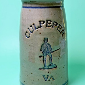 May include: A cylindrical stoneware crock with a speckled beige finish and blue accents. The front features the embossed text CULPEPER above a relief image of a standing figure holding a rifle, with the letters VA positioned below the figure.