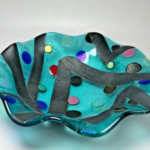May include: A decorative glass bowl with a turquoise base, adorned with black stripes and colorful polka dots in shades of blue, pink, yellow, and green. The bowl has a wavy, irregular rim.